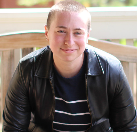 Kendra Albert, a white non-binary person with buzzed hair smiles at the camera. They are wearing a leather jacket.